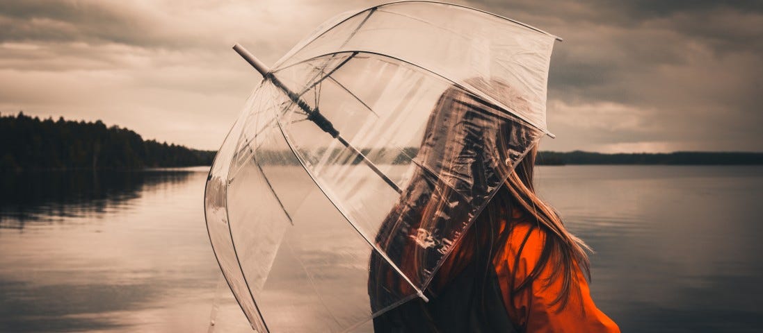 A woman in a red coat stands beside a lake, under a cloudy sky. She holds a clear umbrella.