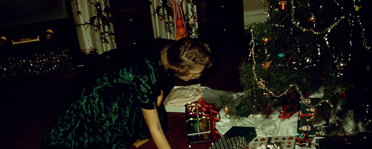 Woman in a green dress kneeling on floor in front of a Christmas tree and packages circa 1950's
