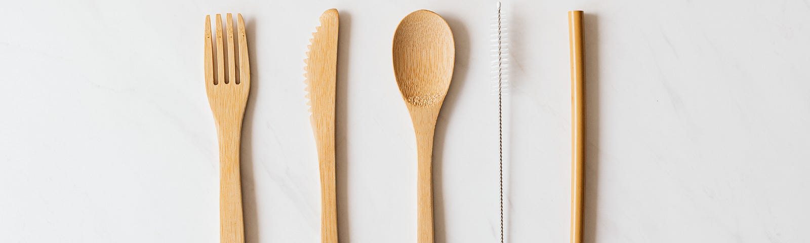 A single place setting of reusable bamboo cutlery on a light grey background. From left to right: a fork, a knife, a spoon, a straw-cleaning brush, and a straw