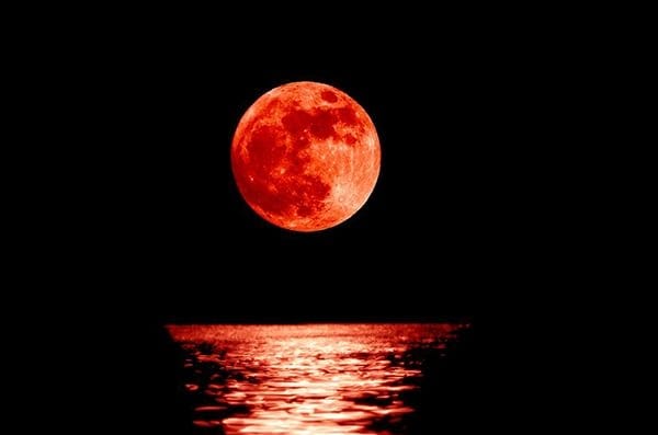 A picture of a blood red moon rising above the water.