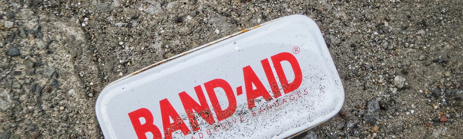 A white metal first aid kit that says BAND-AID in red letters sitting on a patch of dirt