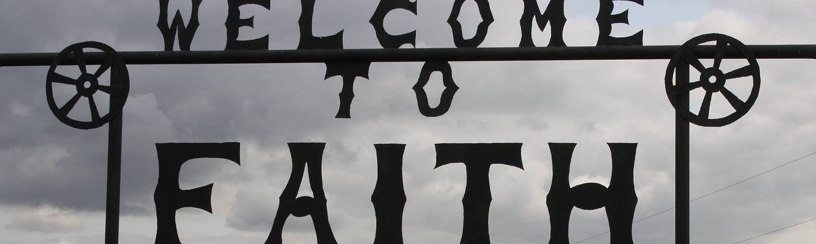 Photo of a black iron sign reading Welcome To Faith EST 1910 against a cloudy sky.