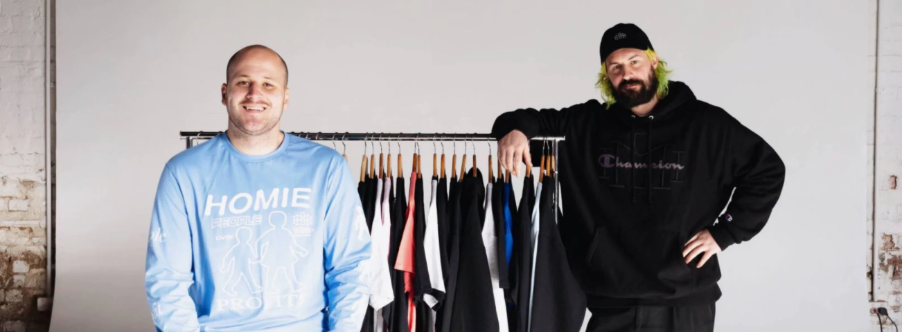 HoMie founders, Nick Pearce and Marcus Cook stand in front of a clothing rack full of t-shirt. Nick is wearing a blue shirt with ‘Homie’ on the front. Marcus is wearing a black jumper and cap.