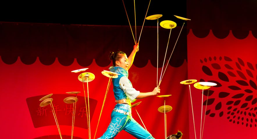 Three young Asian women perform a circus act on stage, holding long poles and spinning plates on them without letting them fall.