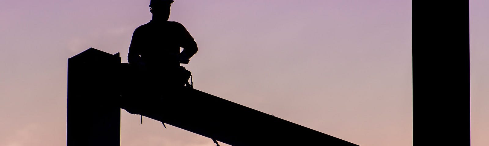 Silhouette of a construction worker sitting  on a high girder against a pink sky.