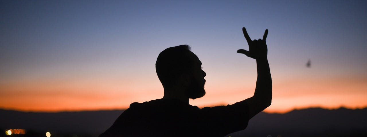 Silhouette of a bearded man in front of a recent sunset, holding his right hand in the I Love You shape.