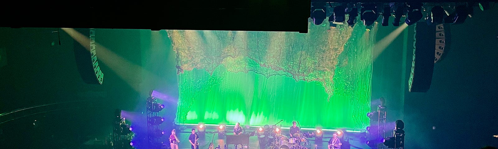 Vance Joy stands at the front of the stage, playing guitar. The band is behind him a saxephone player, a trumpet player, a keyboardist, a drummer and a guitarist. The stage is lit up green and blue.