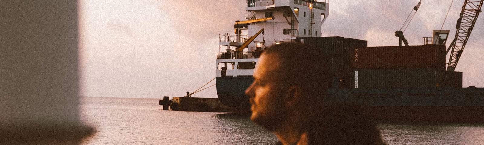 Man and woman sitting on a pier looking at the sunset with a ship in the background.