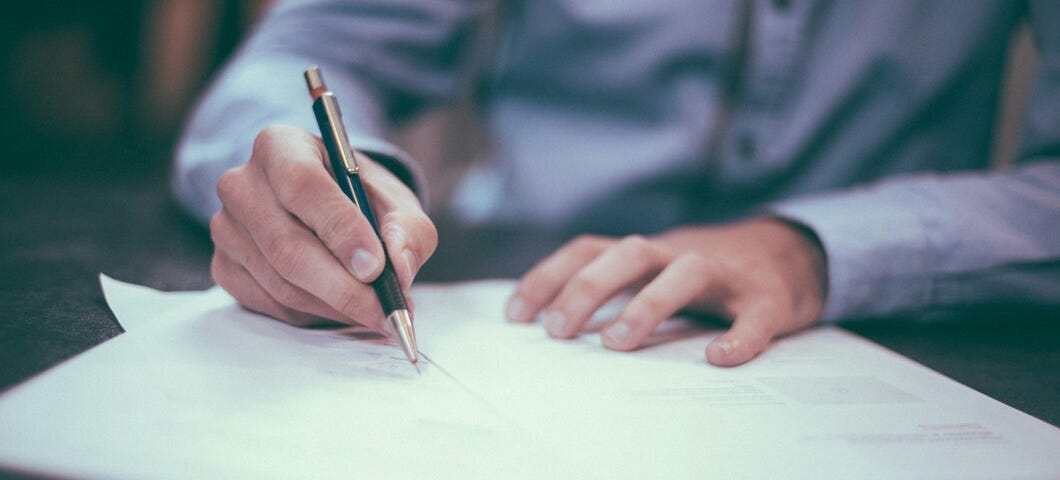 A man’s hand holds a pen. He is signing some documents that are on a table.