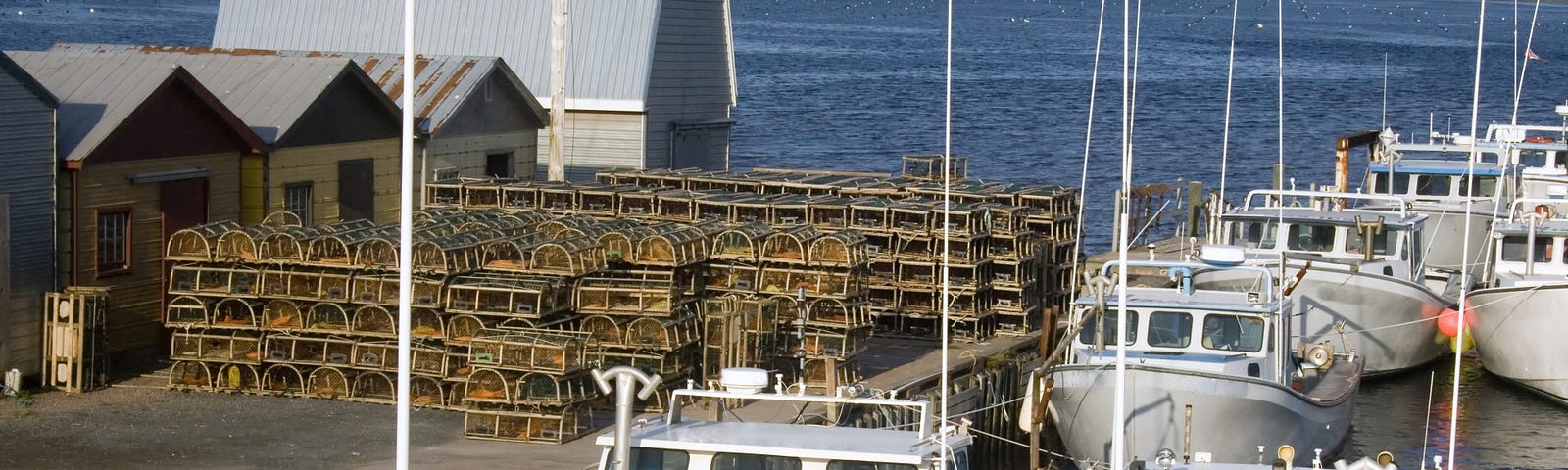 Fishing boats in water along a wharf with lobster traps on the wharf with blue ocean waters in the back and rolling green hills.