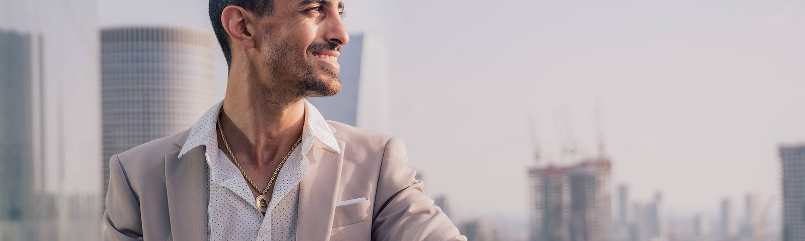 A sharp-dressed man with a handsome, gold watch. He is outside a tall building, leaning with one arm over the balcony and smiling to himself.