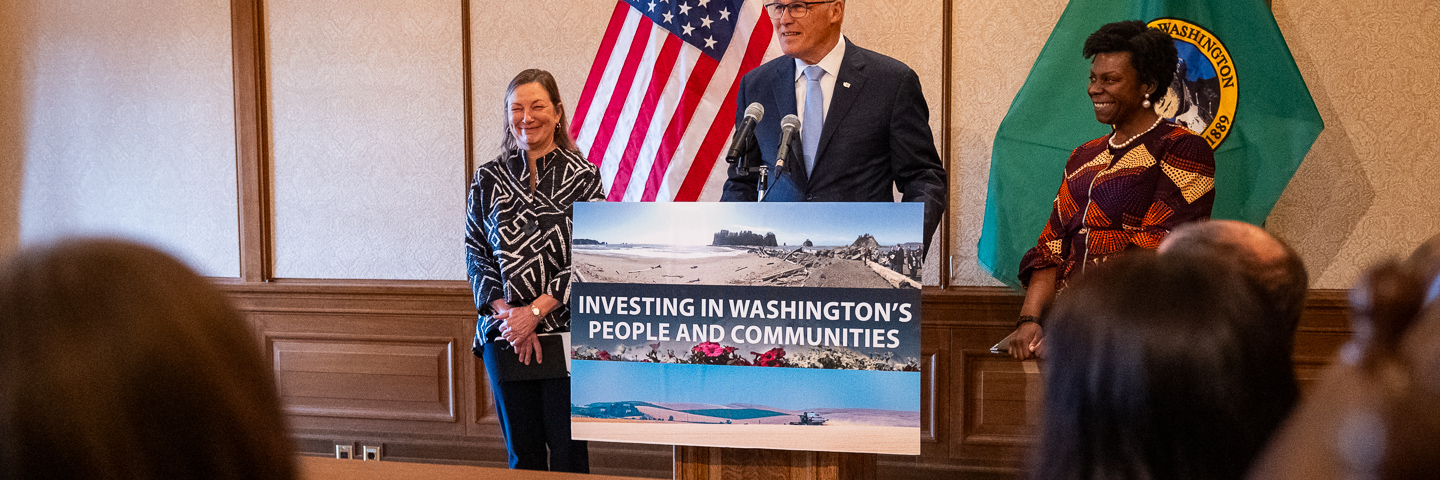 Photo of three people in front of two flags at a podium with a sign: Investing in Washington’s People and Communities