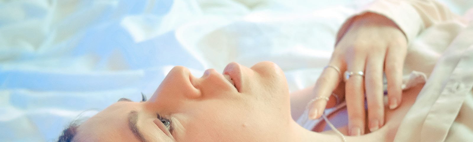 A woman lying on a bed with her hand resting on her chest, gazing upward in soft natural light.