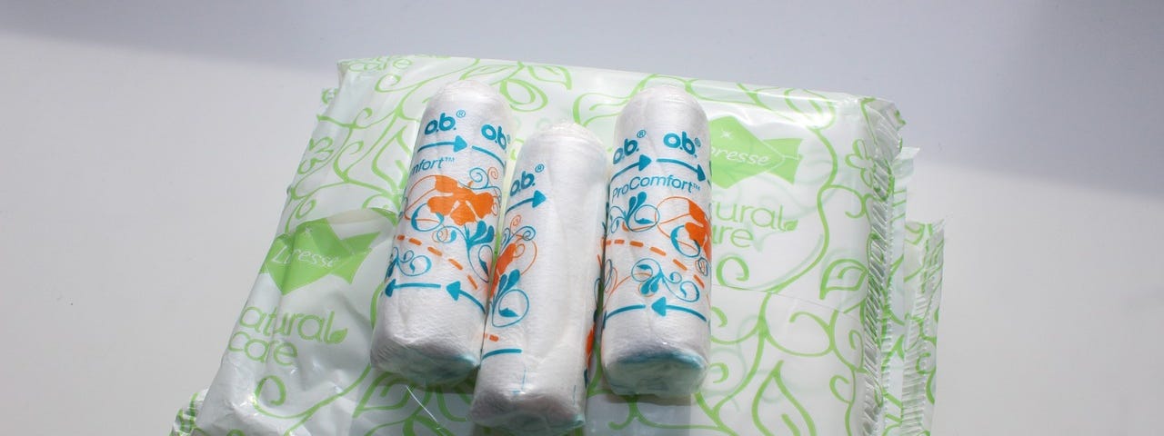 Image shows sanitary pads encased in white plastic with green lines, and on top of that three tampons in their plastic encasings.