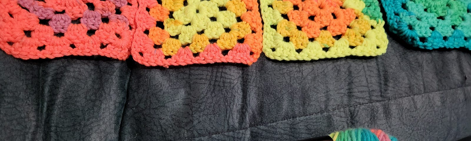 4 multicoloured crocheted granny squares, arranged across the back of a grey couch, ranging in colour from purple to orange to yellow to green to blue. Below them is a skein of variegated yarn in the same colours.