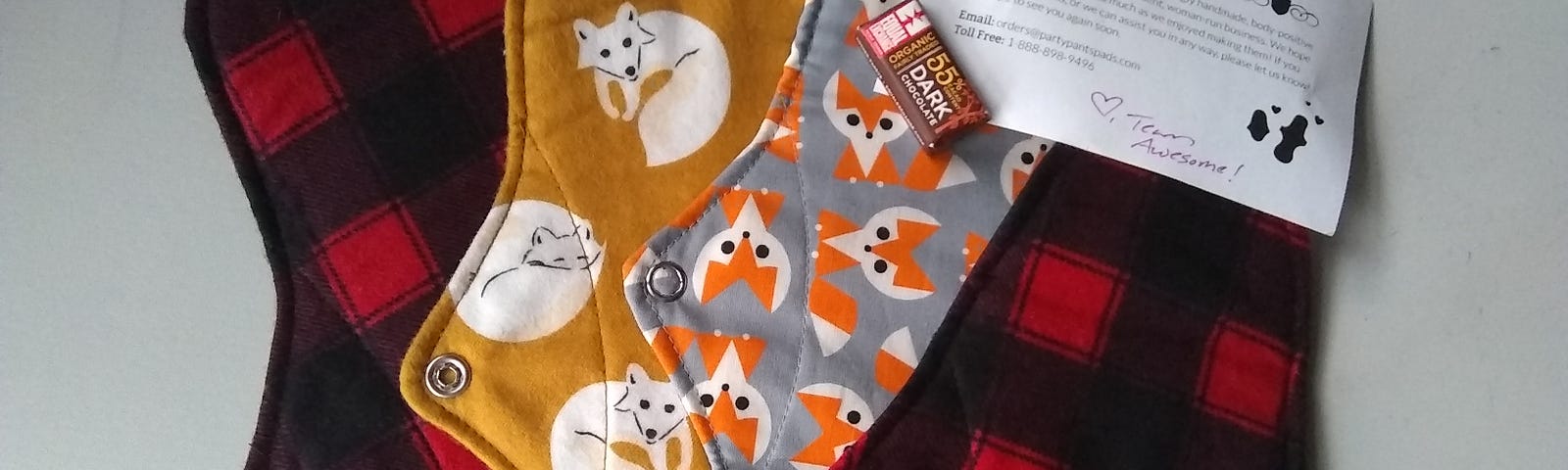 Four cloth menstrual pads. Two of them are red and black plaid, one of them has white foxes on a gold background, and one has orange and white foxes on a grey background.