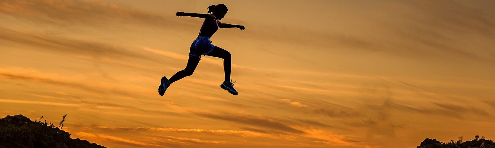 Woman jumping high between two hills, silhouetted against a sunset.