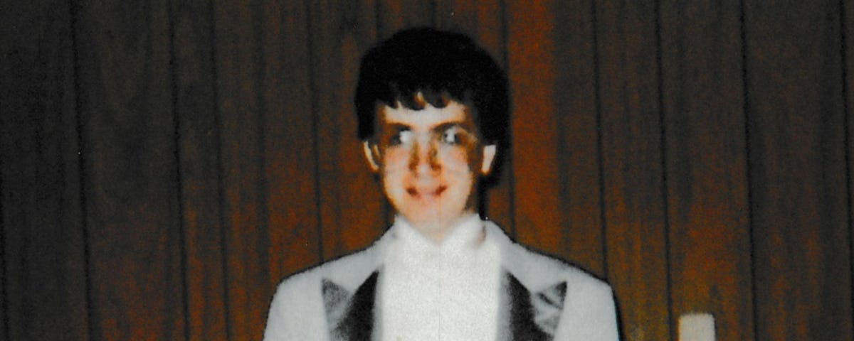 Young man wearing a grey dinner jacket with wide darker lapels and a white shirt. He is wearing glasses and is standing in front of a wood panelled wall.