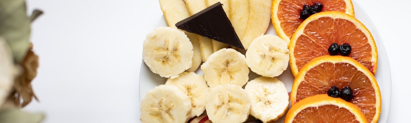 A round plate on a white background containing slices of orange with blueberries on top, slices of apple, sliced of banana, and two triangles of dark chocolate. There are some white flowers on the left.