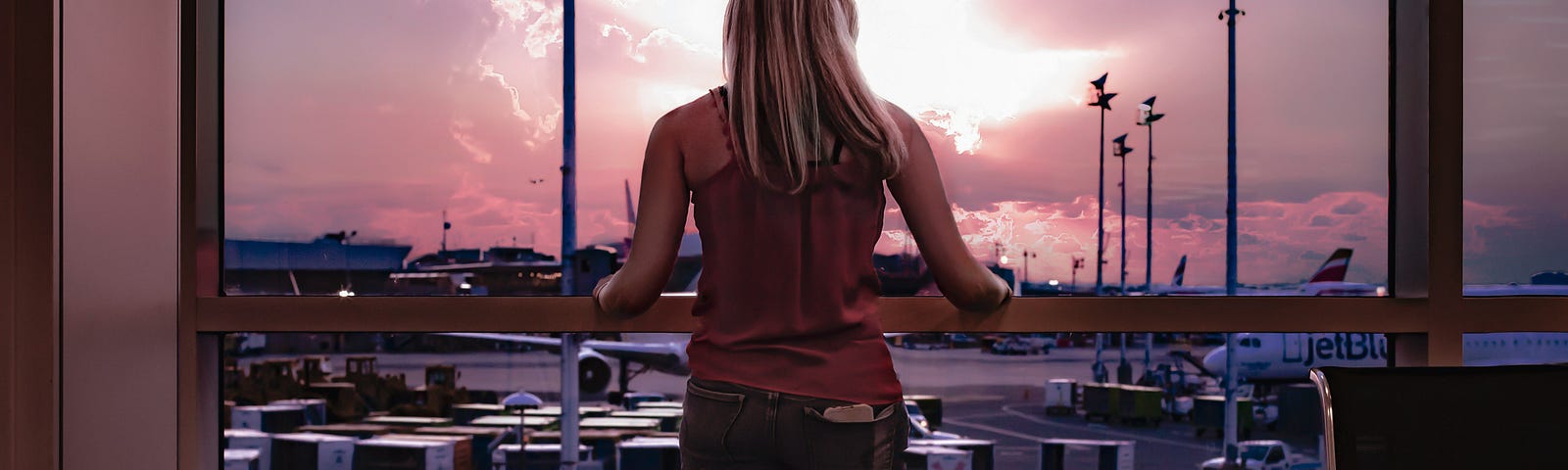 A blonde woman wearing jeans and a red singlet top stares out an airport window that overlooks the tarmac. The sky looks ominous as the sun peeks through the clouds. It’s a forlorn setting. Contemplating her destiny perhaps.