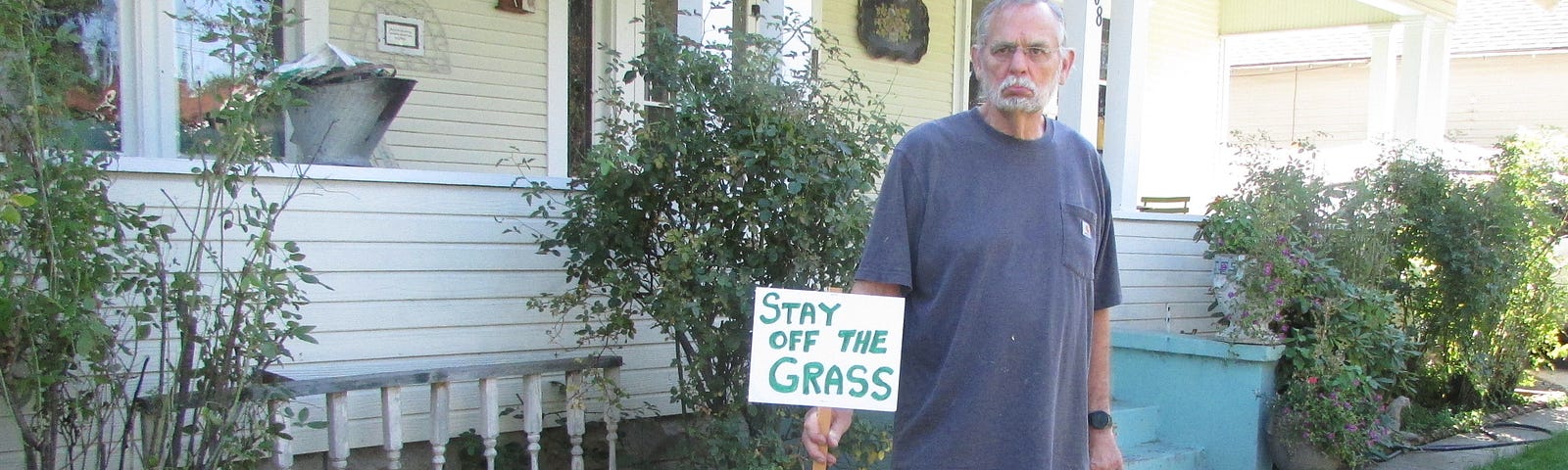 grumpy old man holing keep off the grass sign in front of house with dog