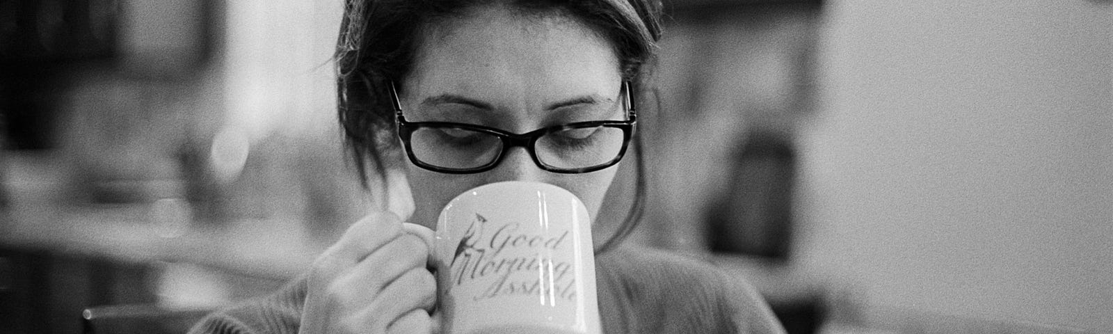 Black and white photo of a woman with her hair up, wearing glasses, drinking out of a mug that says “good morning asshole” on it