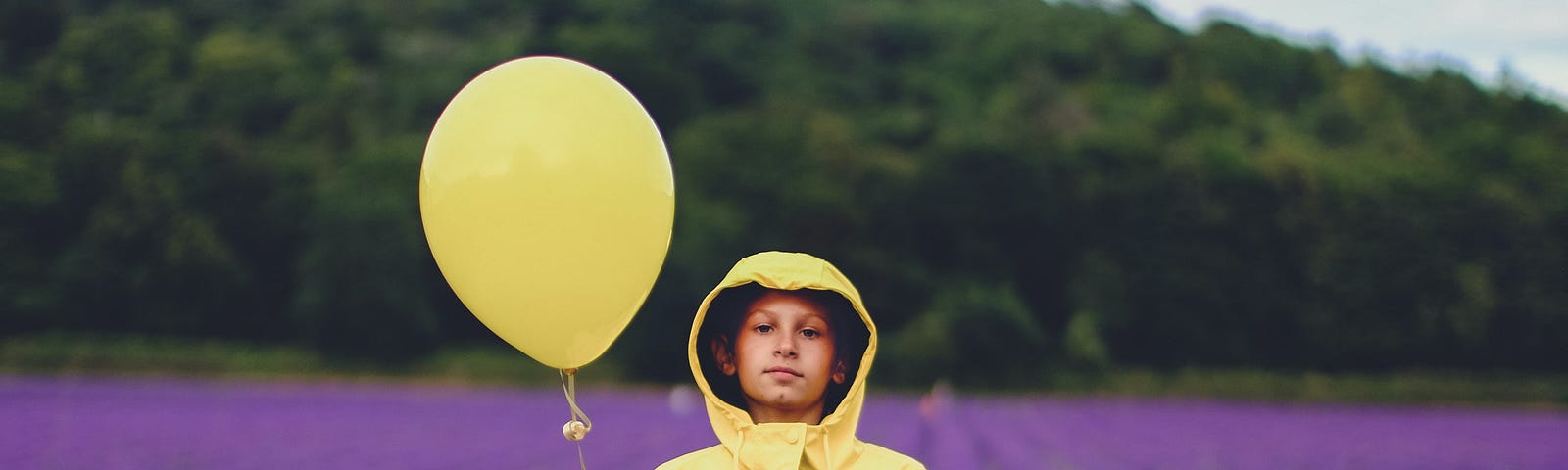 A woman standing in a filed of purple lavender in a yellow rain coat hiolding a yellow balloon.