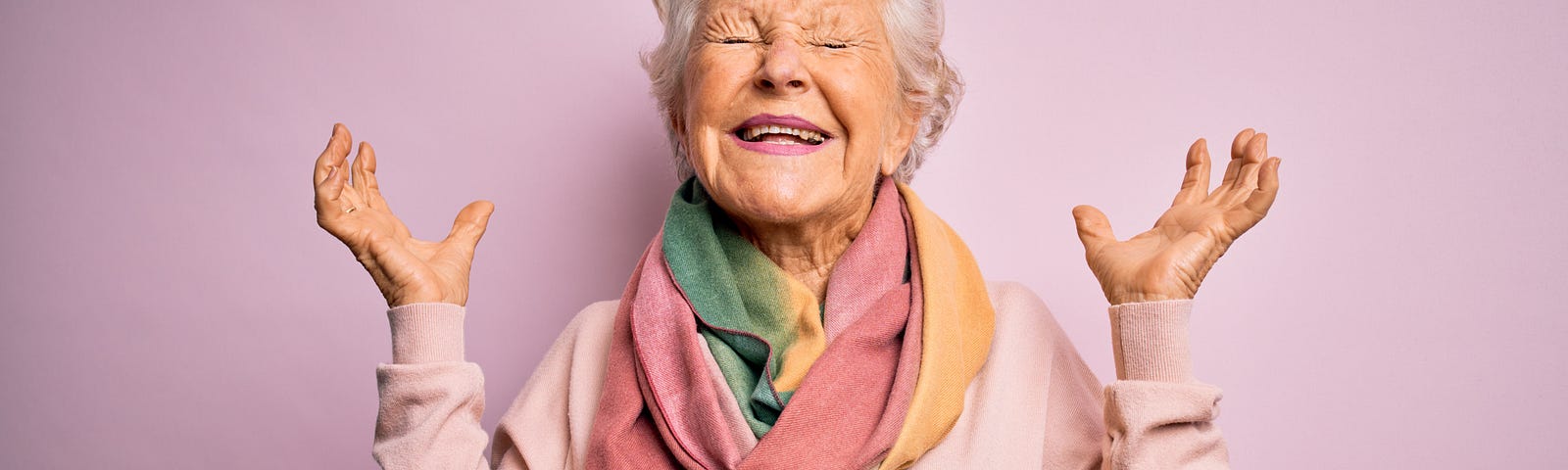 Senior lady on pink background, eyes closed with arms up in excitement