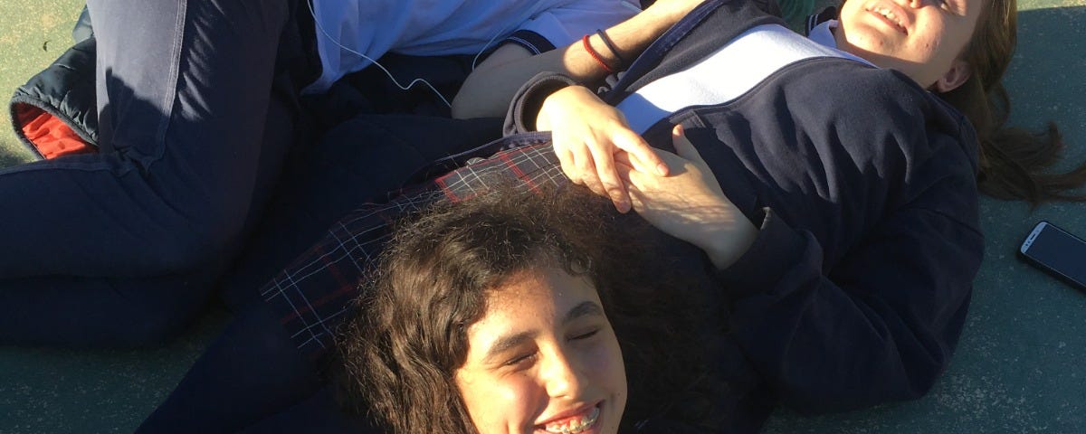 Gianna acostada en el patio de su escuela junto a dos amigues, sonriendo bajo el sol.