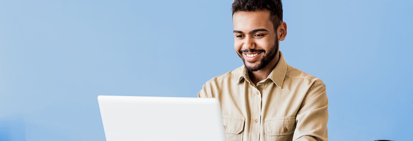 A young man smiling while attending a remote learning experience through a laptop with a blue wall in the background.