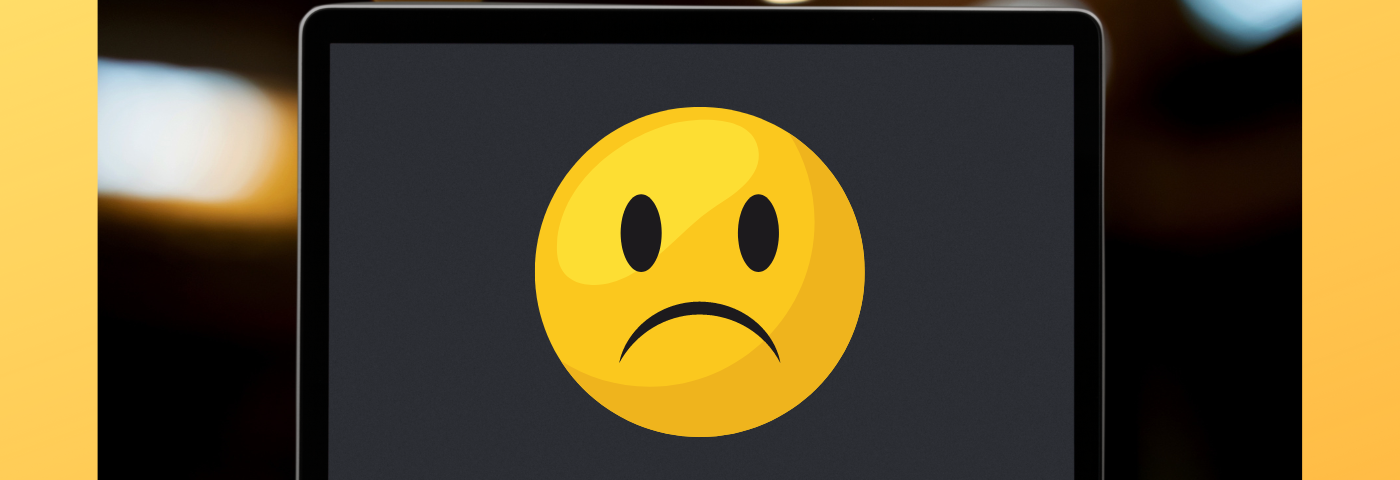 A laptop computer sits on a wooden desk and shows a sad emoji face.