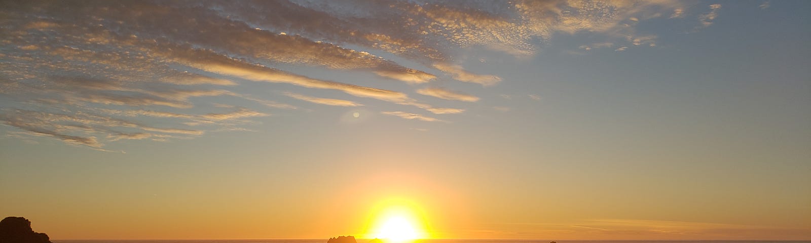 Wide angle photograph of a man from behind who’s facing the setting gold ball of the sun over Pacific ocean waves, with faint clouds overhead along the California coast.