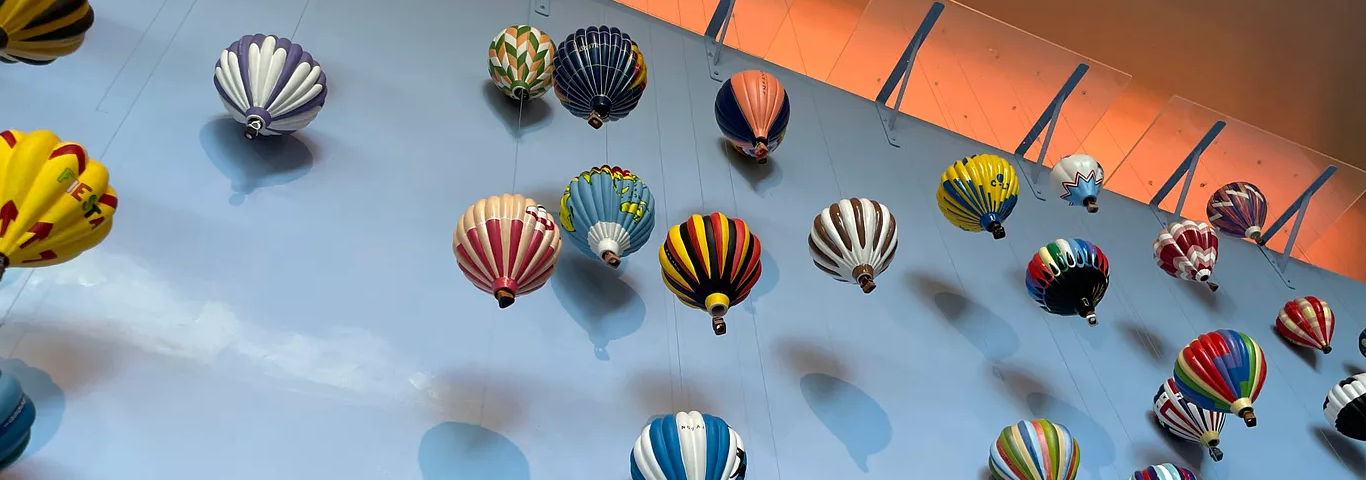 Various miniature hot air balloons, shown from below. They are mostly striped and multicolored, and hang against a light blue wall.