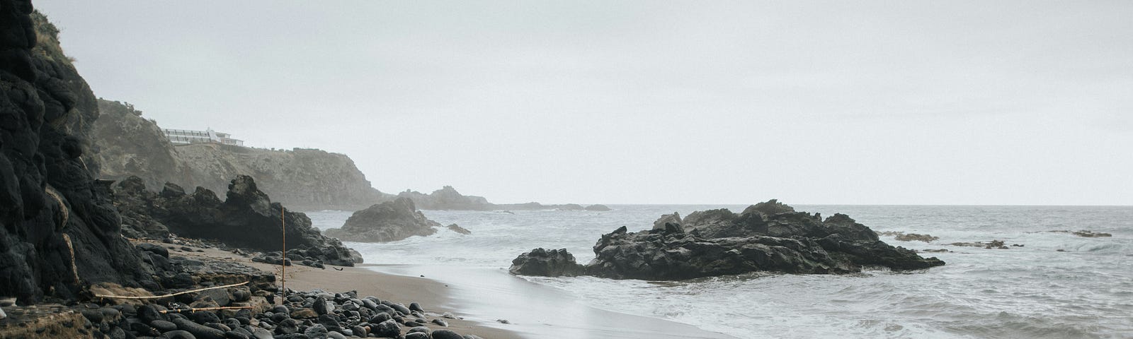 Photograph of a beach with dark-coloured sand and rocks on an overcast day.