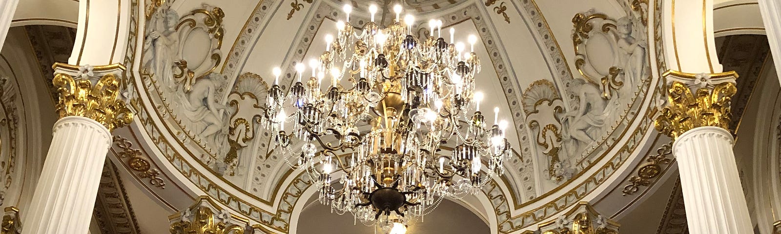 Grand hall with pillars and a chandelier hanging from a gilded ceiling