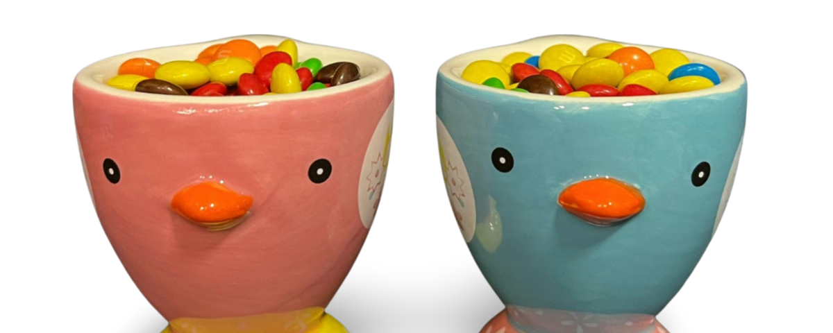 Two novelty eggcups in the shape of baby chicks. Each has been filled with mini M&Ms. The baby chick eggcup on the left is pink and the one on the right is blue. White background.