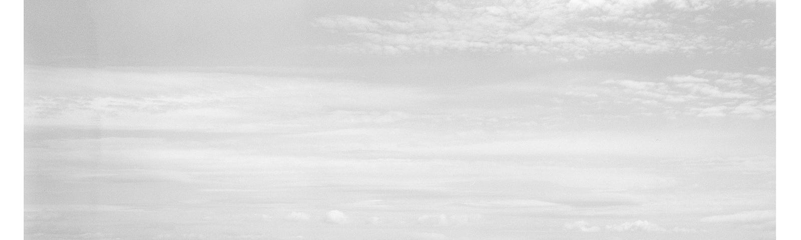 The image is a black-and-white photograph of a serene beach scene. The foreground features a flat, sandy shoreline with subtle wave patterns. The midground shows the ocean, with gentle waves rolling in. The horizon line is straight, separating the sea from the sky, which is mostly clear with a few scattered clouds. The overall atmosphere is calm and peaceful, with minimal elements, emphasizing the vastness of the sea and sky.