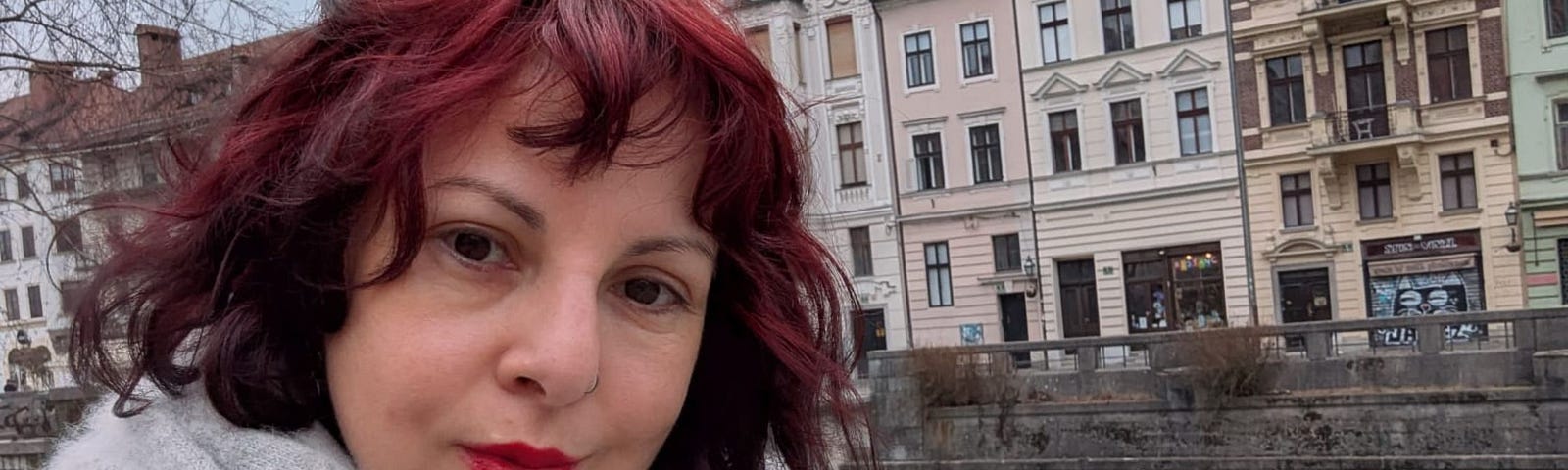 Sarah Jayne, a white woman with brightly dyed read hair looks into the camera. Behind here is a river and tall terraced houses in central Ljubljana.