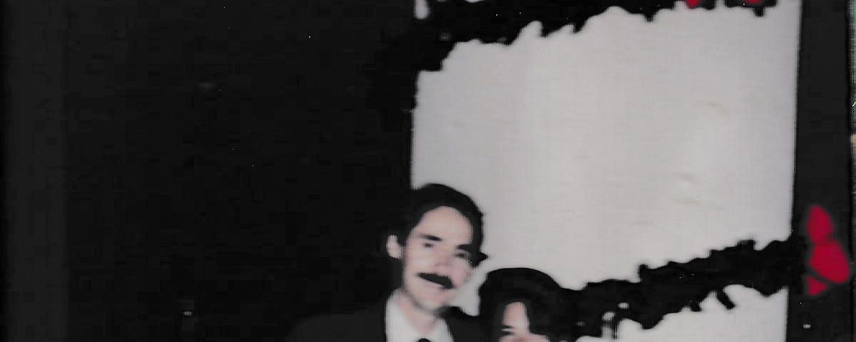 Photo of John and I (author) at Elton John concert sponsored by Yamaha where he worked. John is wearing a suit and tie, and has a mustache and dark hair. His arm is around me and I am leaningvinto his shoulder, my left arm touching his suit by his chest. I am wearing a black formal dress with trim. We are standing in front of a large white pole around which were draped a few thick strands of greenery with large red-pink flowers at their sides.