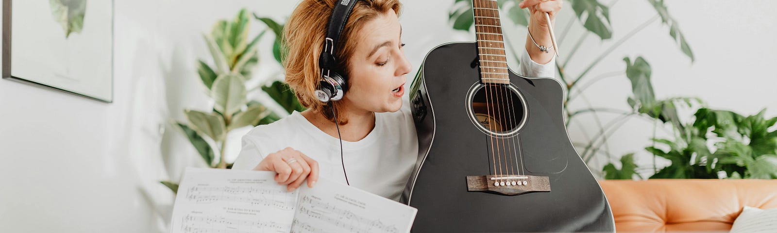 Woman Playing Guitar on Video Call on Computer. She is wearing a headset. She is pointing at the strings and has some sheet music in her right hand.