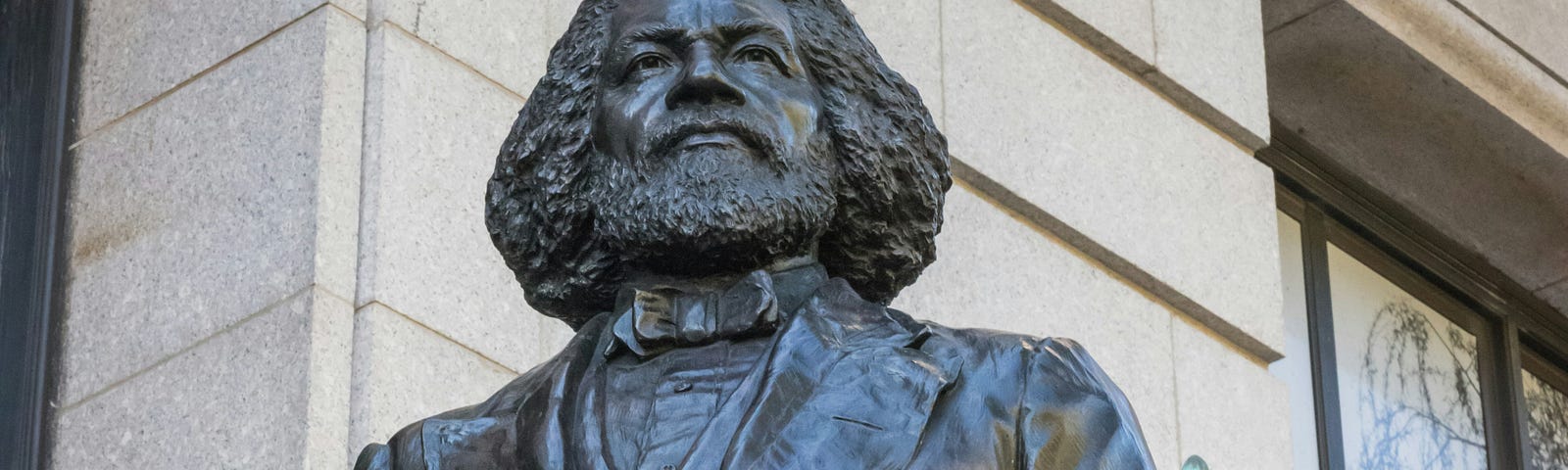 A photo of the Frederick Douglass Statue at the New York Historical Society, standing tall, proud and holding a book in his left hand close to his body.