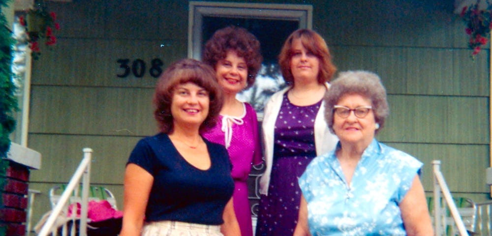 Four women are standing on the steps in front of a house, smiling at the camera. They are dressed in varied and colorful outfits, suggesting a cheerful and possibly familial gathering.