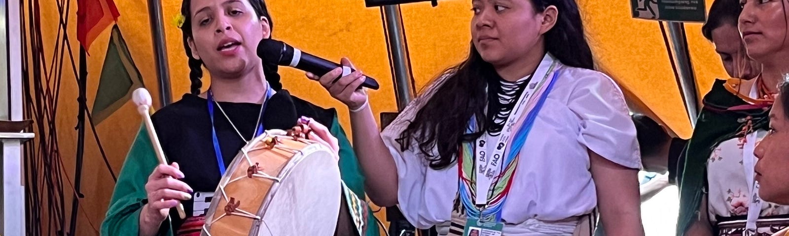 A person holding and playing a tambor drum and singing. Another person next to them is holding a microphone up to capture their performance