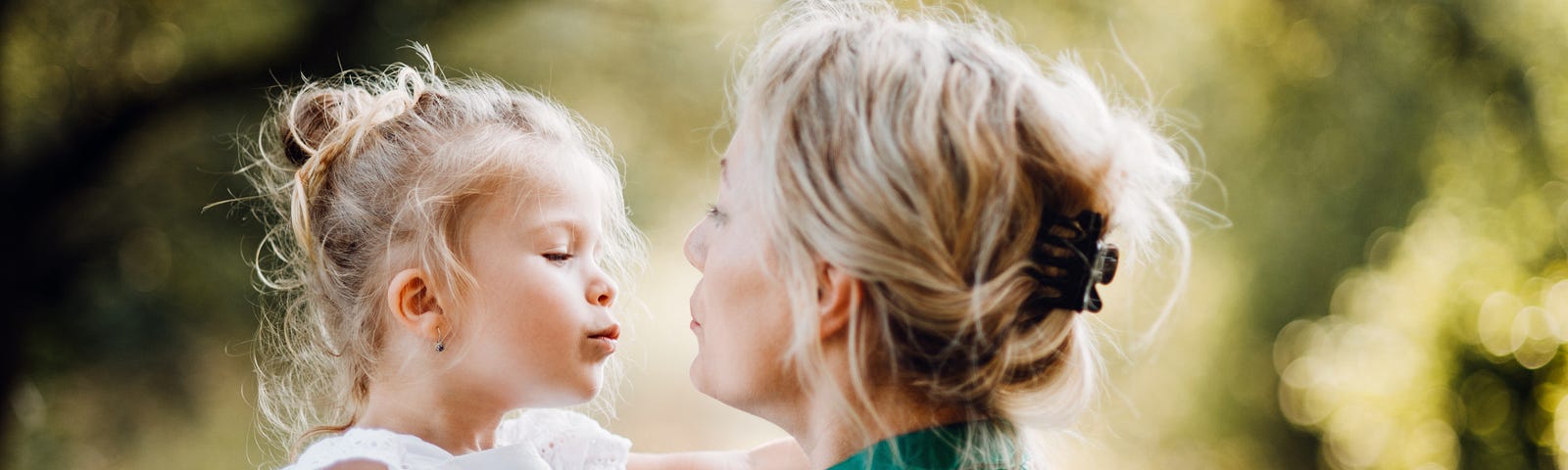 A blonde mother and daughter embrace one another under the shade of summer trees.