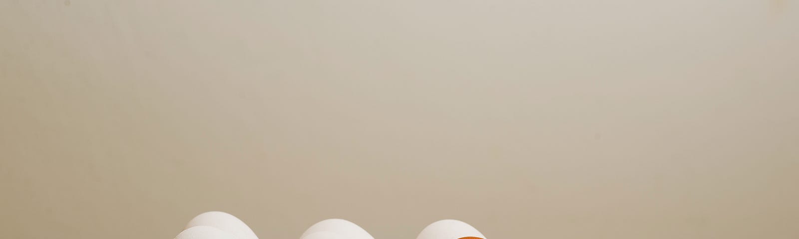 There are six eggs sitting in a egg carton made of glass. Five of them are white, one of them is orange. All of the eggs have cartoon faces. The white eggs look angry at the orange egg, and the orange egg looks scared.