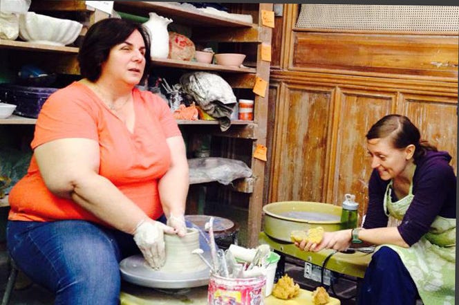 Michelle, left, works with clay on a pottery wheel. Another woman, right, wearing a green apron, smiles at Michelle’s work.