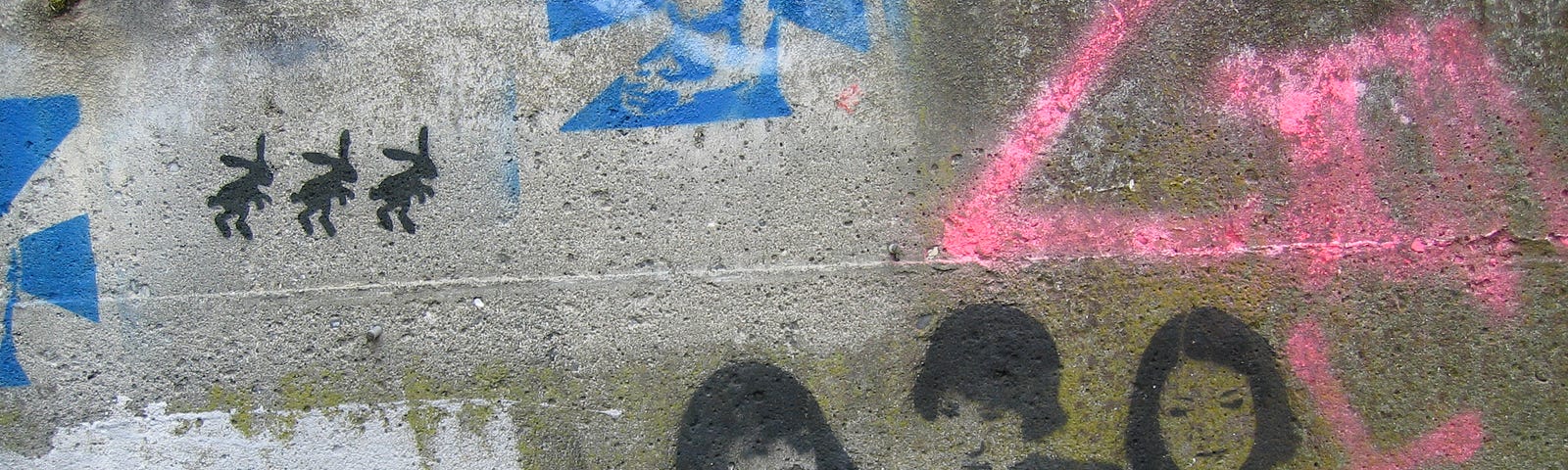 A photo by Moira Cloney, showing green, mouldy concrete covered with graffiti, including a stencil of Sleater-Kinney’s faces.