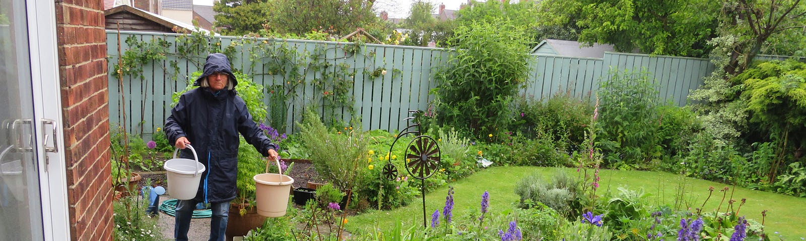 Linda Acaster wearing waterproofs, bucketing water around a green garden  during a downpour May 2024.