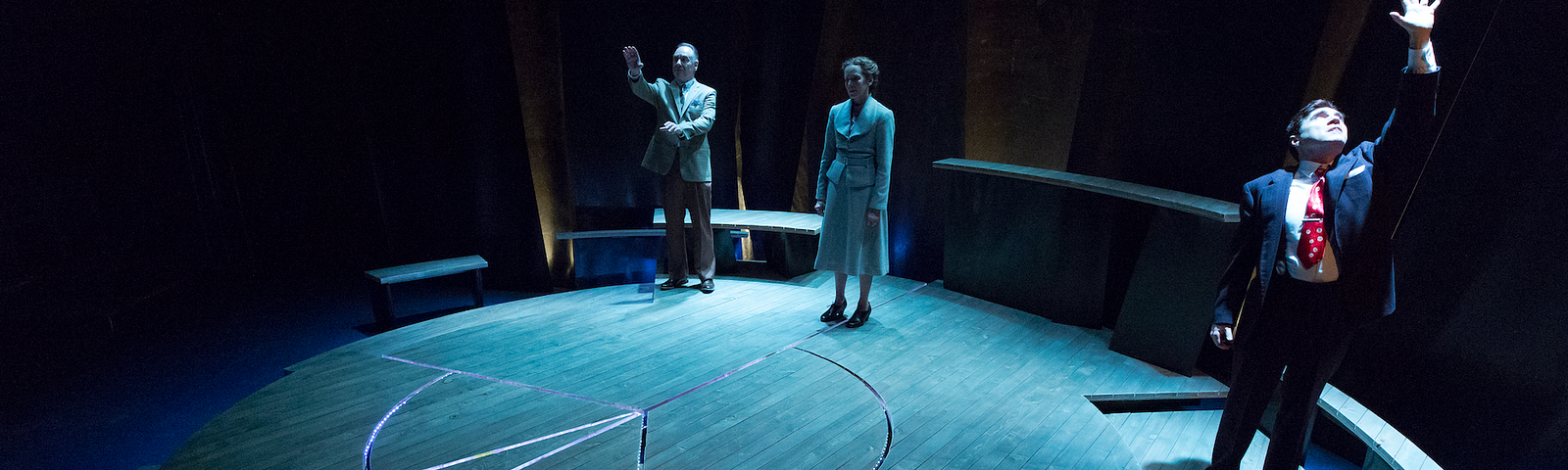 Three people in 1940s clothing stand on a stage with a circular platform in blue light