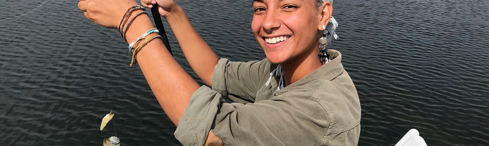 Young person of color smiling, sitting on a dock holding a fishing line with a fish she caught on the hook